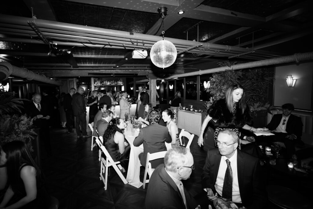 Black and white photo of guests seated and dining under disco ball at wedding reception