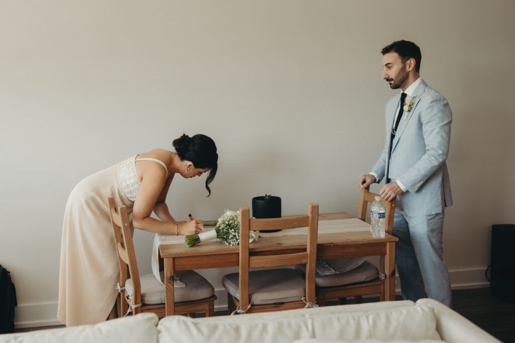 Bride signing paperwork at a dining table while the groom stands nearby in their apartment