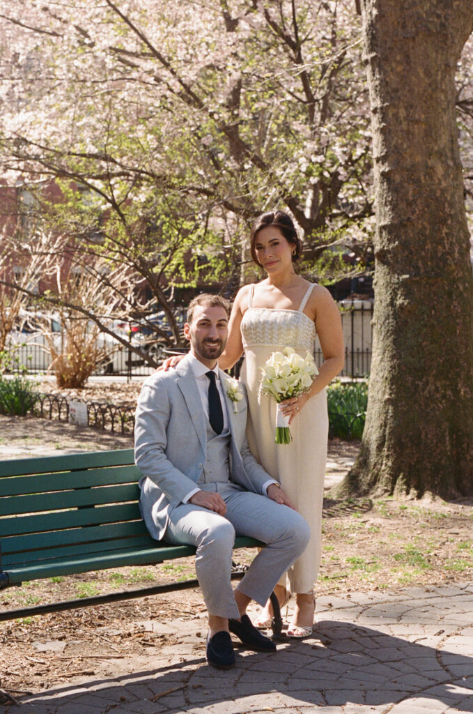 Bride standing beside seated groom on a green park bench during spring wedding portraits