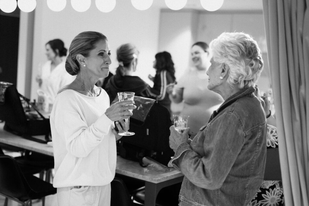 Two women holding drinks and chatting during wedding morning preparations in a softly lit room