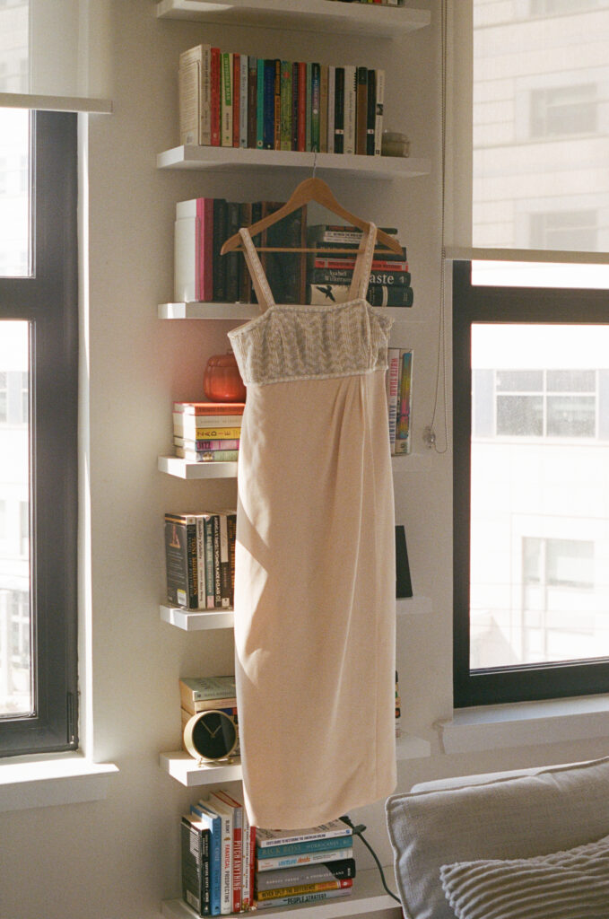Minimal wedding dress hanging in front of bookshelves and large apartment windows with sunlight streaming in