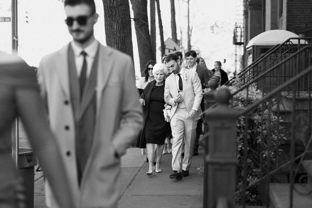 Black and white candid of wedding guests walking along a city sidewalk toward the reception venue in Jersey City