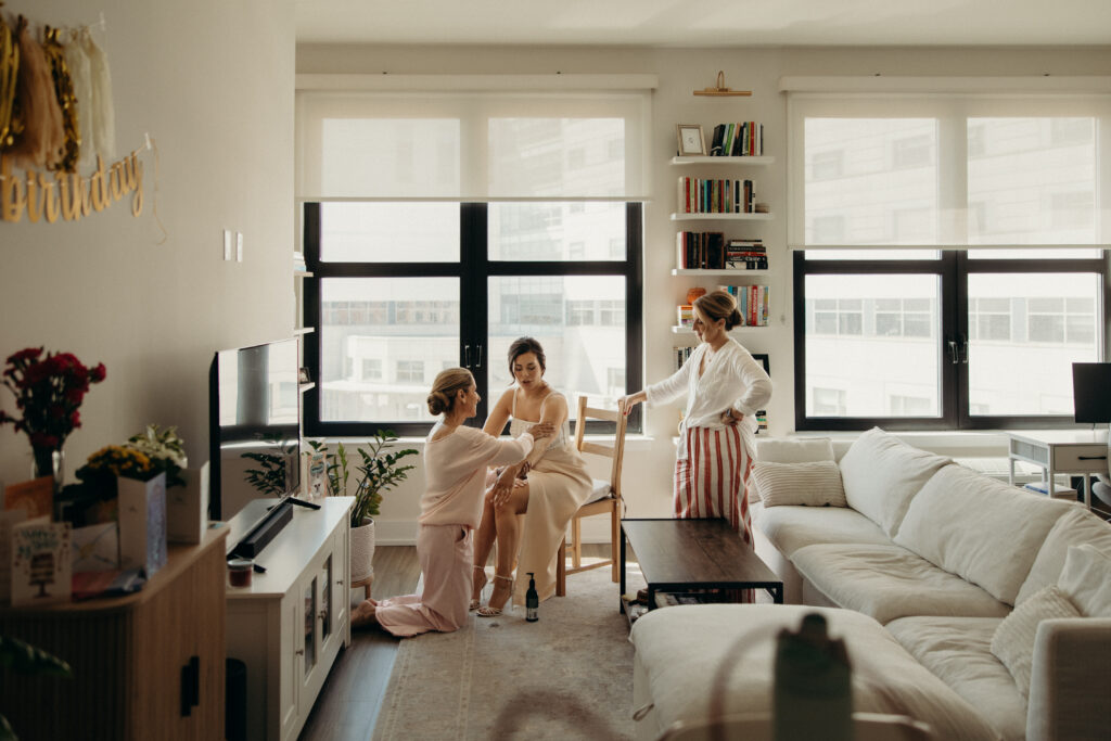 Bride sitting while family members help adjust her dress in a bright modern apartment