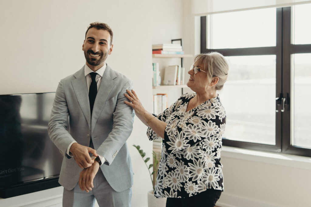 Groom smiling while an older woman adjusts his light blue suit jacket before the wedding