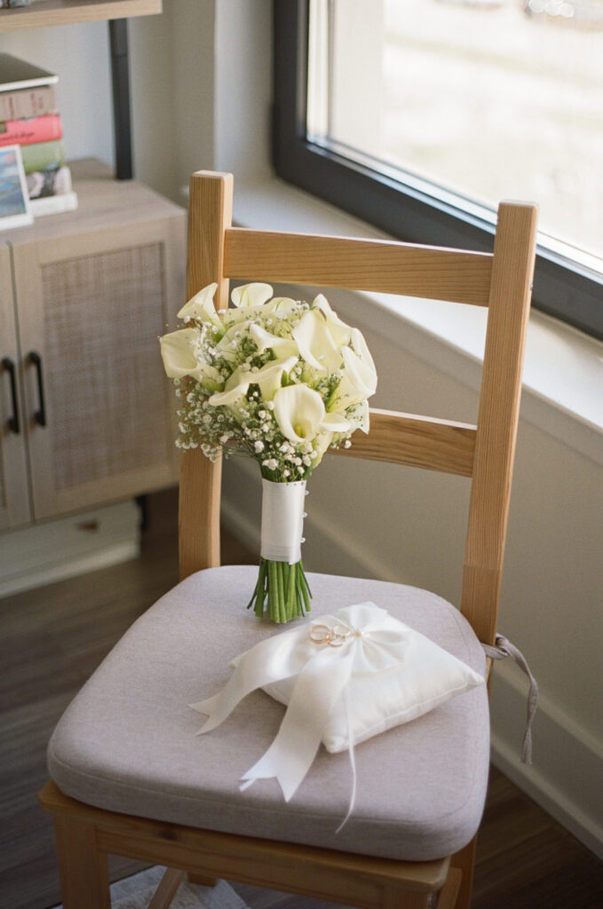 White calla lily bouquet and wedding rings displayed on a wooden chair beside a bright window