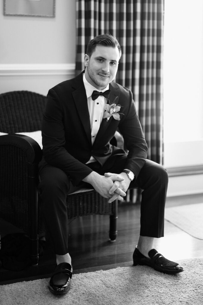 Groom seated in a chair wearing a black tuxedo before the ceremony at The Ashford Estate