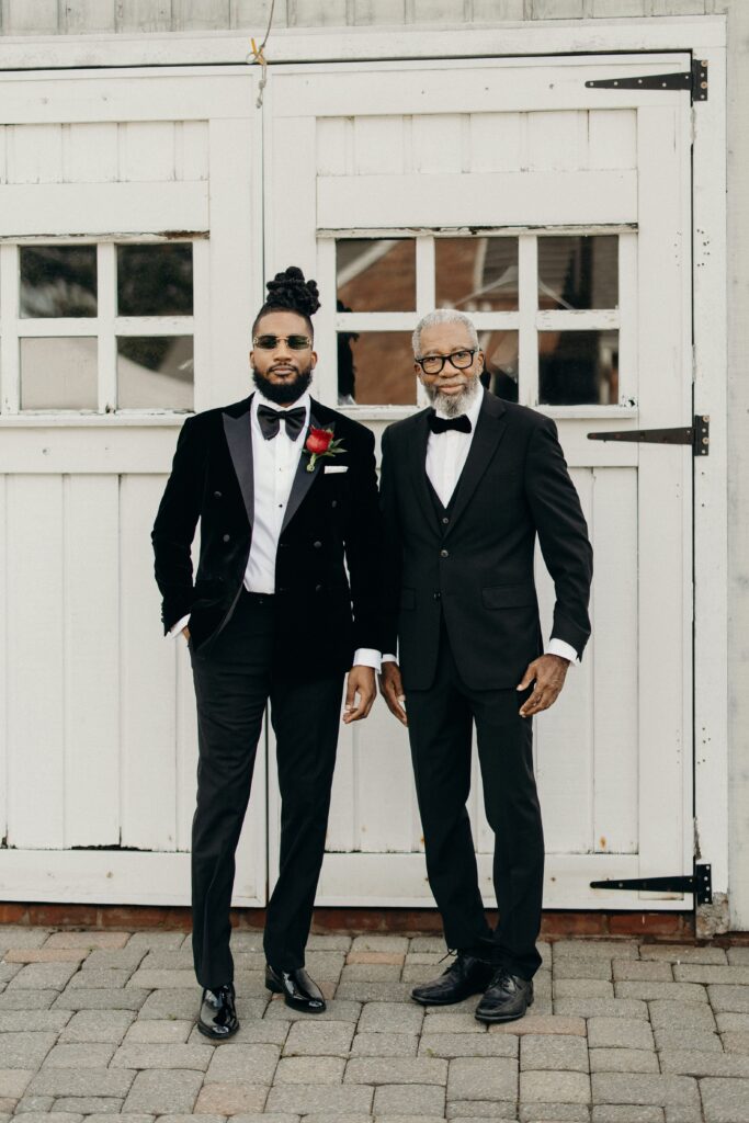 Groom posing with a guest in black suit attire in front of a white barn door at Perona Farms