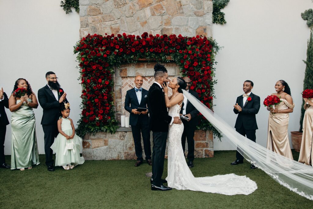 Bride and groom kissing at the altar under a red rose archway at Perona Farms