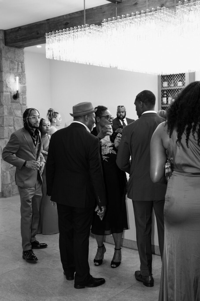 Black and white shot of wedding guests mingling during cocktail hour under a modern chandelier at Perona Farms