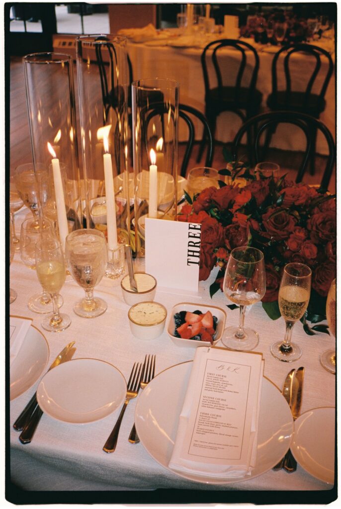 Close-up view of an elegant reception table setup with tall candle cylinders, red floral centrepieces, and printed menus at Perona Farms