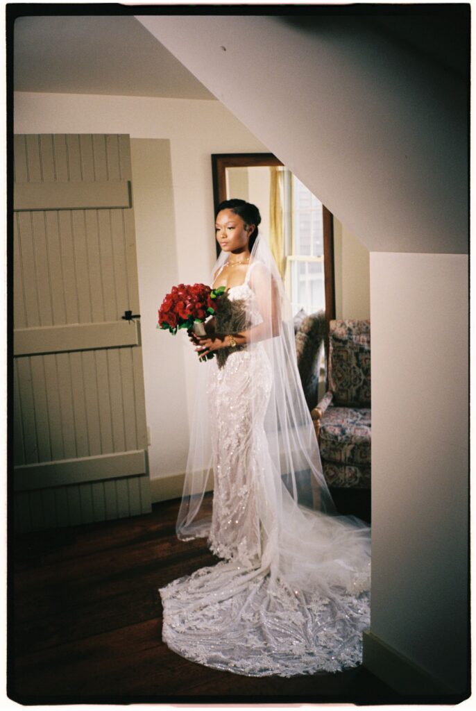 Bride standing in a white lace dress and long veil holding a bouquet of red roses at Perona Farms