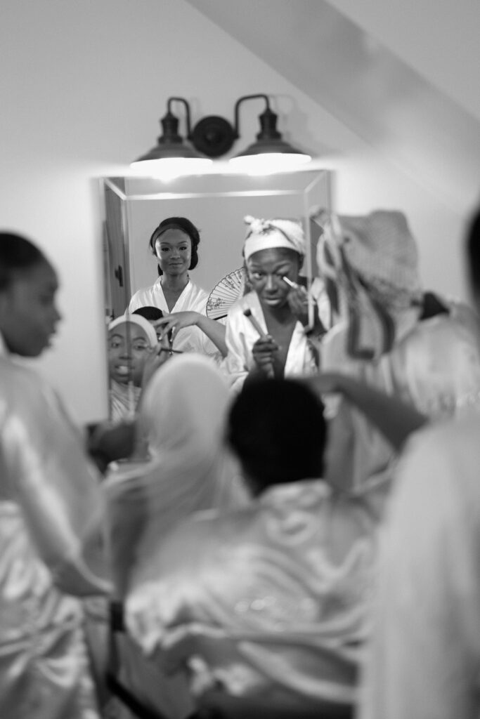 Black and white candid of bridesmaids getting makeup done in front of a mirror at Perona Farms