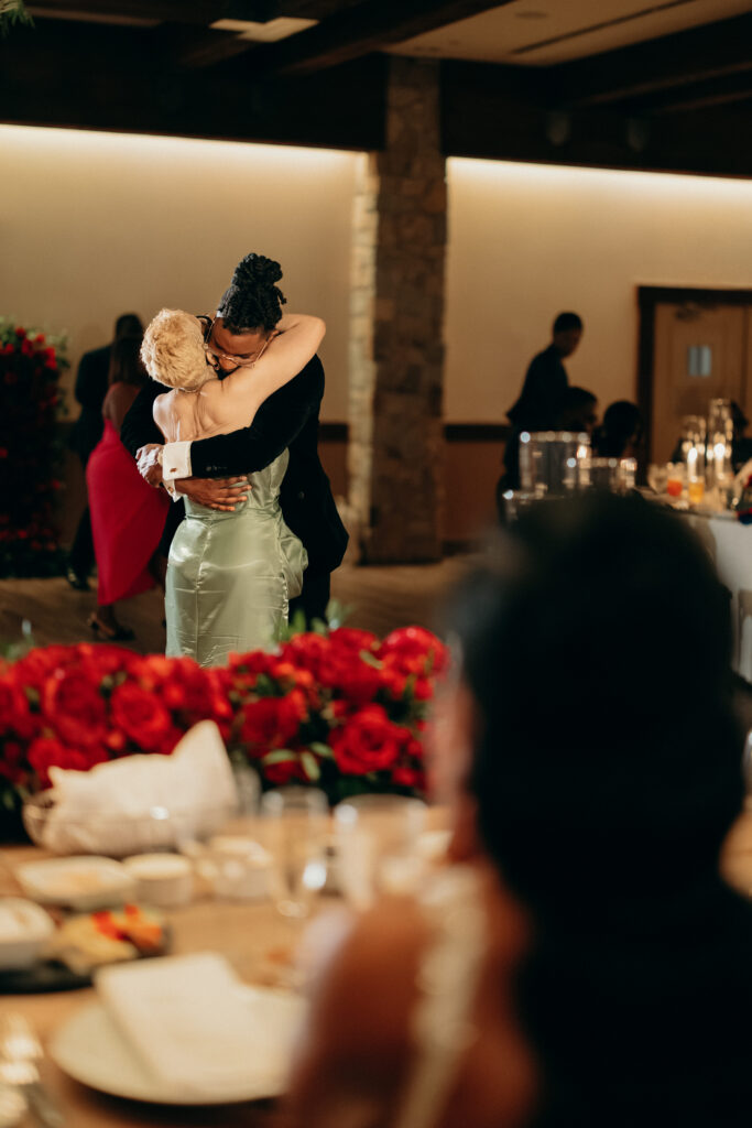 Groom embracing a guest on the dance floor surrounded by red floral arrangements at Perona Farms