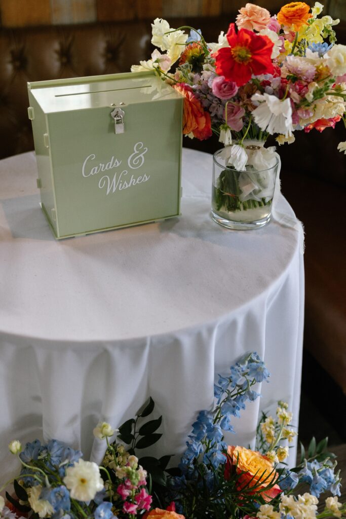 Green cards and wishes box with floral arrangements on a white-clothed table at Battello