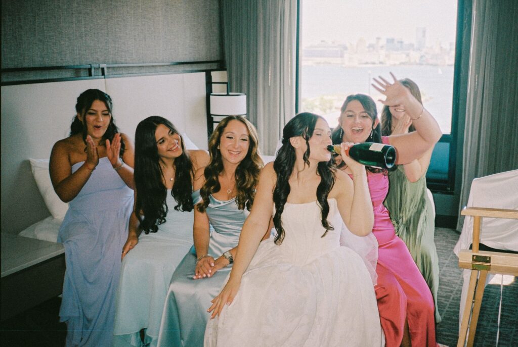Bride drinking champagne straight from the bottle surrounded by her bridesmaids in a hotel room before heading to Battello