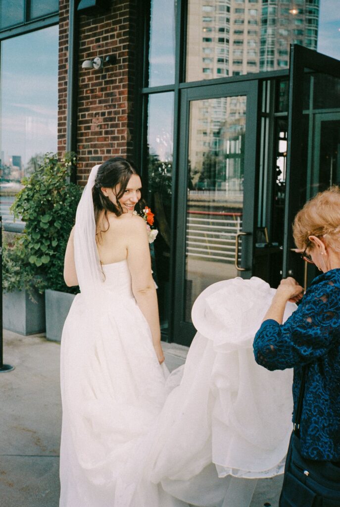 Bride looking back over her shoulder as someone adjusts her wedding dress train outside Battello